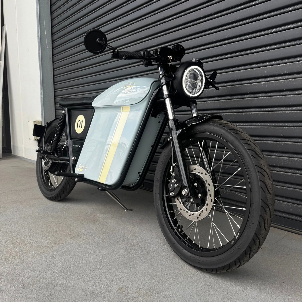 A Streetdog electric motorcycle parked in front of a garage roller door. The tank and side panels are baby blue and feature a cream stripe.