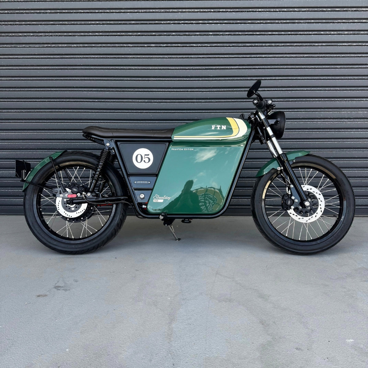 A limited edition phantom Streetdog electric motorcycle parked in front of a garage roller door. The tank and side panels are dark green with light cream and white accents.