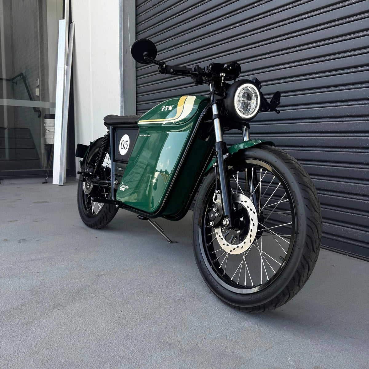 A limited edition phantom Streetdog electric motorcycle parked in front of a garage roller door. The tank and side panels are dark green with light cream and white accents.