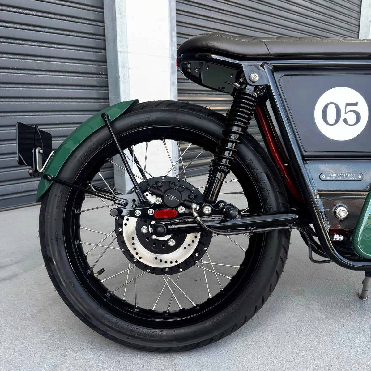 A limited edition phantom Streetdog electric motorcycle parked in front of a garage roller door. The tank and side panels are dark green with light cream and white accents.