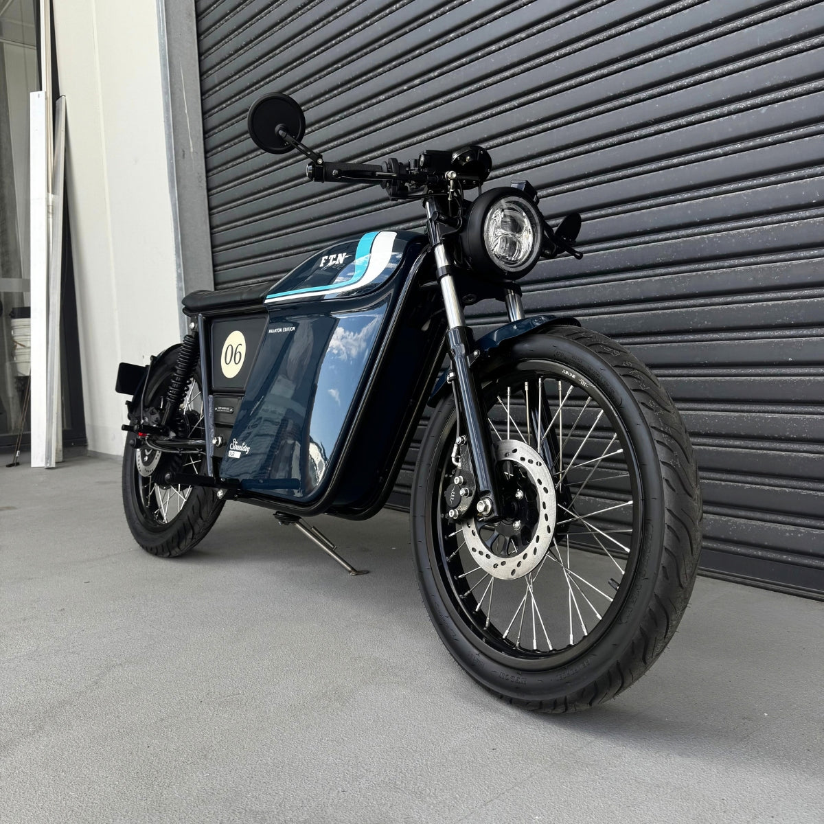 A limited edition phantom Streetdog electric motorcycle parked in front of a garage roller door. The tank and side panels are dark blue with light blue and white accents. 