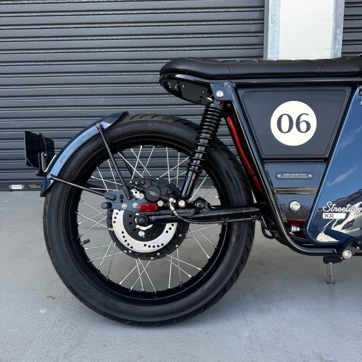 A limited edition phantom Streetdog electric motorcycle parked in front of a garage roller door. The tank and side panels are dark blue with light blue and white accents. 